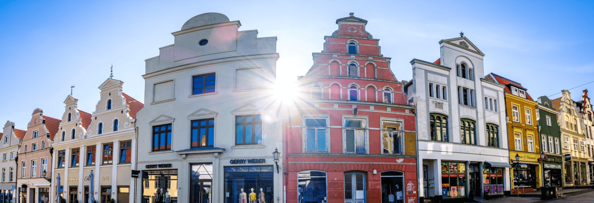 Farbenfrohe historische Giebelhäuser in einer Häuserzeile bei strahlendem Sonnenschein mit markantem Lichtstern-Effekt.