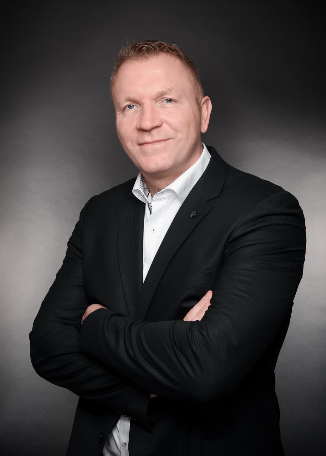 Porträt eines lächelnden Mannes im schwarzen Sakko und weißem Hemd mit verschränkten Armen.